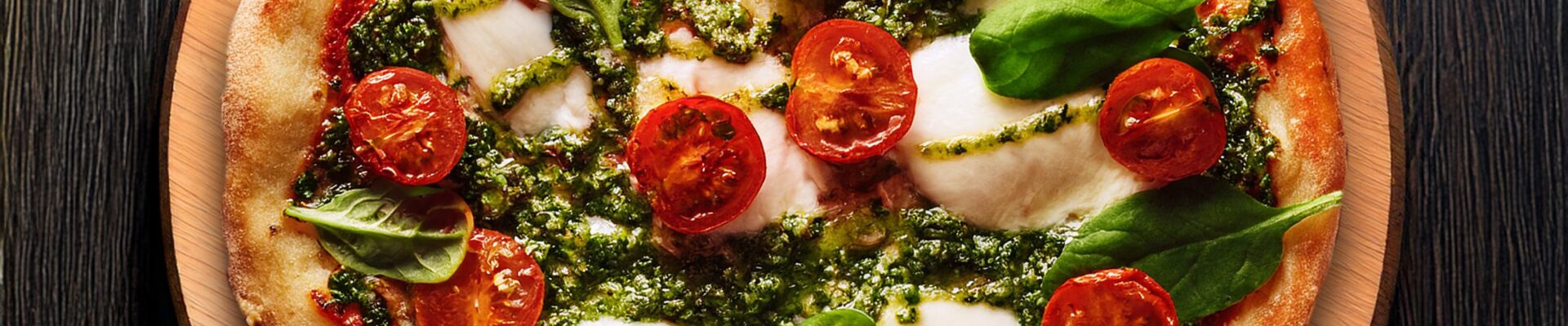 A freshly baked pesto pizza made on a lebanese flat bread base, topped with bocconcini, cherry tomatoes, and fresh basil leaves on a wooden board.