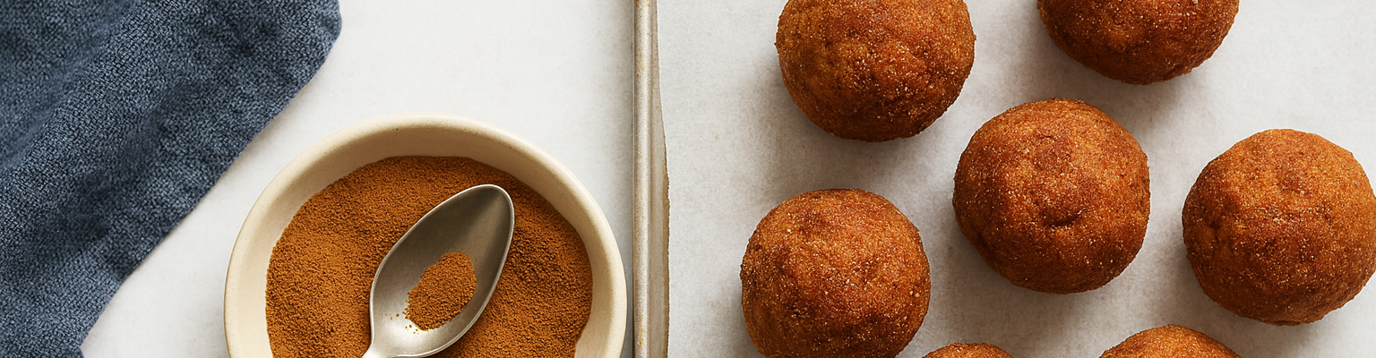 Golden-brown cinnamon balls on a baking tray with a bowl of ground cinnamon and a spoon.