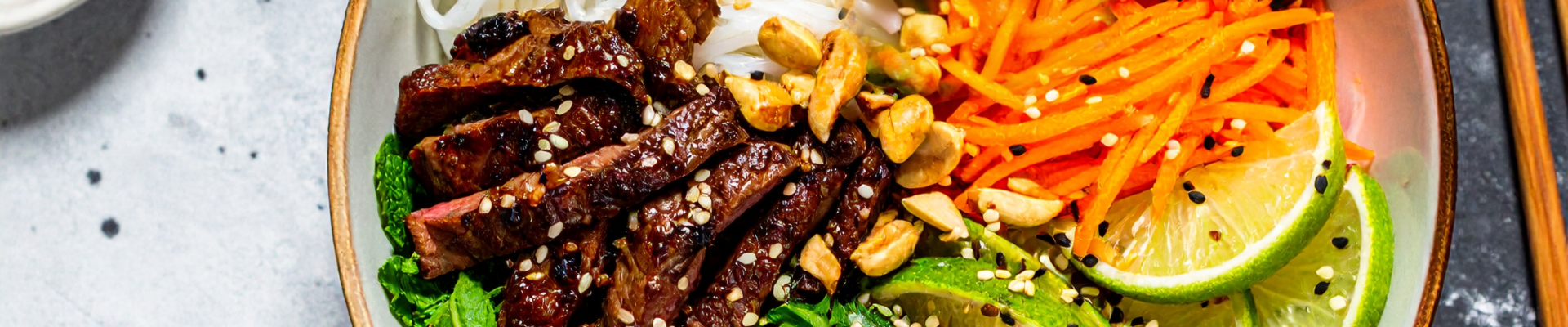 Grilled beef vermicelli bowl with fresh herbs, shredded carrots, lime wedges, and sliced chili.