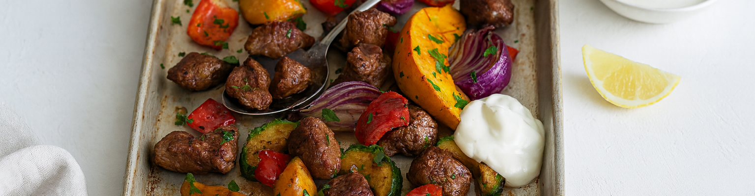 Tray bake of roasted meat and vegetables with sweet potato, zucchini, red onion, and capsicum, served with yoghurt and lemon wedge.