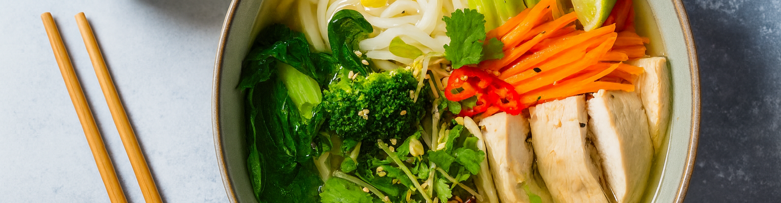 Chicken pho noodle soup with fresh herbs, bok choy, broccoli, carrots, and lime in a clear broth.