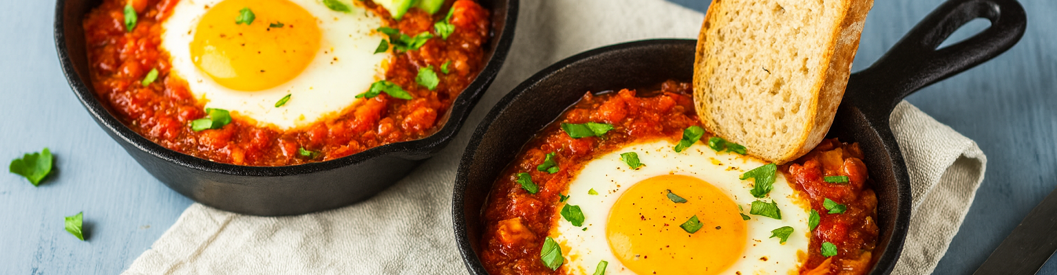 Shakshuka in cast iron skillets with sunny-side-up eggs, tomato sauce, avocado slices, and toasted bread.