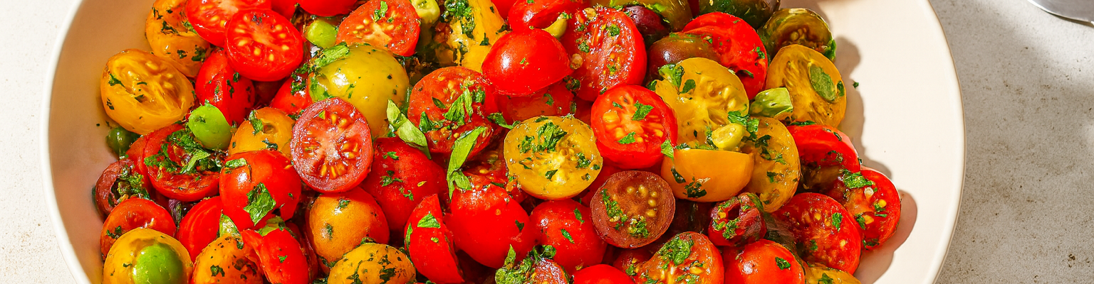 Tomato and olive salad with chopped herbs in a white serving bowl with a wooden spoon.