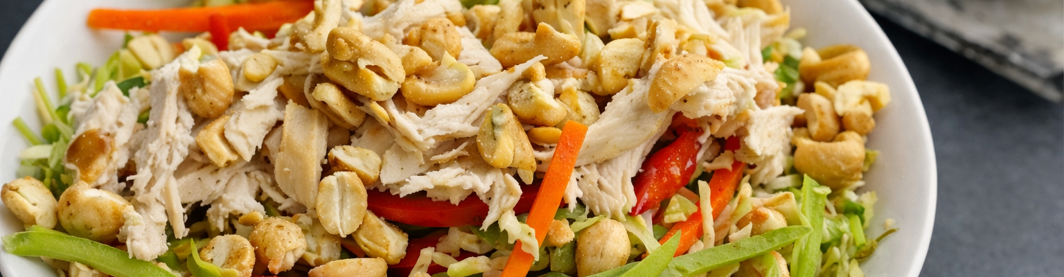 A bowl of chicken noodle salad with shredded chicken, sliced vegetables and roasted cashews.