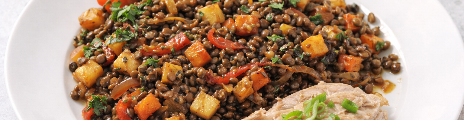 A white plate holding a warm lentil and vegetable salad with diced root vegetables and red capsicum, served with a piece of fish garnished with sliced green shallots.
